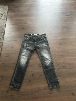 Replay - Säljer ett par grå jeans med slitningar och tvättade detaljer. Jeansen har raka ben, fem fickor och klassisk knappgylf. Perfekta för en avslappnad och trendig look. Använda 1/2 gånger.