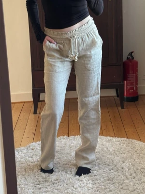 Beiga linnebyxor med dragsko - Beiga linnebyxor med elastisk midja med snören att dra åt. Byxorna har raka ben och är tillverkade i linnematerial. Köpta för 800kr från Skagen Clothing förra året, aldrig använt. Storlek XS. 