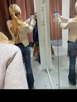 Beige stickad tröja med öppen rygg - Trendig beige stickad tröja med öppen rygg och dekorativa knytband. Tröjan har långa ärmar och en något croppad passform. Perfekt för dig som vill ha en unik och snygg detalj på din outfit. Materialet känns mjukt och bekvämt.