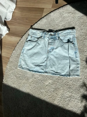 Ljusblå jeanskjol - Snygg ljusblå jeanskjol  i klassisk modell med fickor. Kjolen har en kort passform och stängs med knappar som dragkedja framtill. Perfekt att matcha med en t-shirt eller hoodie för en chill look. Perfekt nu till sommaren 