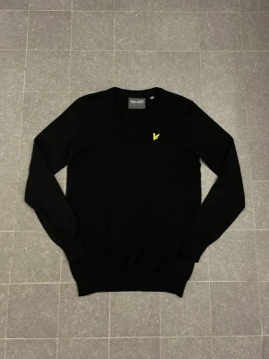 Svart stickad tröja från Lyle & Scott - Klassisk svart stickad tröja från Lyle & Scott med V-ringning och den ikoniska gula örnloggan broderad på bröstet. Tröjan har långa ärmar och ribbade muddar vid ärmslut och nederkant. Perfekt till lager på lager och enkel att matcha.