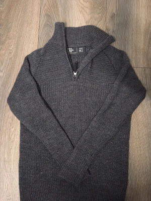 Mörkgrå stickad tröja Neo Mondo - Mysig mörkgrå stickad tröja från Neo Mondo med halv dragkedja vid halsen och ribbade muddar. Perfekt för kyliga dagar och enkel att matcha med andra plagg. Tröjan har en klassisk rak passform och är tillverkad i mjukt material.