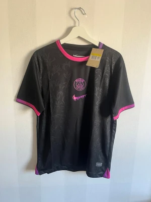 Svart PSG x Nike t-shirt med rosa detaljer - Snygg svart PSG t-shirt från Nike med diskret blommönster i tyget och rosa detaljer vid krage och ärmslut. PSG-logga och Nike swoosh i rosa framtill, samt trycket 'GTO' i nacken. Perfekt för träning eller streetwear.