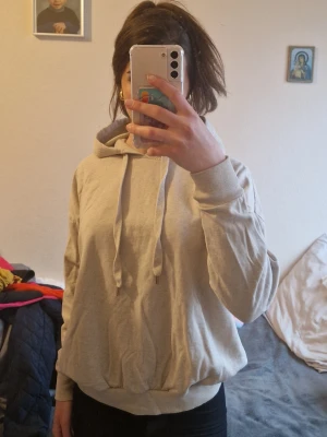 Beige hoodie med huva och dragsko - En basic beige hoodie med huva och dragsko. Hoodien har långa ärmar, ribbade muddar vid ärmslut och nederkant samt en avslappnad passform. Perfekt för en chill och enkel stil. Materialet känns mjukt och bekvämt, troligen bomullsblandning. Inga synliga loggor eller tryck.