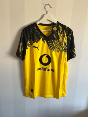 Borussia Dortmund gul matchtröja Puma - Säljer en gul och svart Borussia Dortmund matchtröja från Puma med coolt grafiskt mönster på axlarna. Tröjan har klubbens logga och Vodafone-tryck på framsidan samt DORTMUND på ryggen. Perfekt för fotbollsfans som vill sticka ut på läktaren.