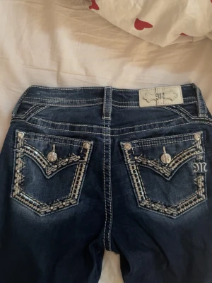 Mörkblå Miss Me jeans med bling - Säljer ett par mörkblå jeans från Miss Me med snygga detaljer i form av bling och broderier på fickorna. Jeansen har kontrastsömmar, låg midja och dekorativa knappar. Lite slitage vid hälarna. Därav priset❣️