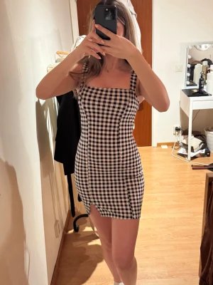 Rutig svartvit kort klänning - Supersnygg kort klänning med svartvitt rutmönster och breda axelband. Klänningen har en tight passform och en liten slits framtill. 