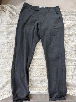 Mörkgrå slim fit kostymbyxor - Snygga mörkgrå kostymbyxor i slim fit-modell med klassiska fickor och bälteshällor. Byxorna har en modern, smal passform och är perfekta för en stilren look. Materialet är mjukt och har en lätt glans som ger en exklusiv känsla.