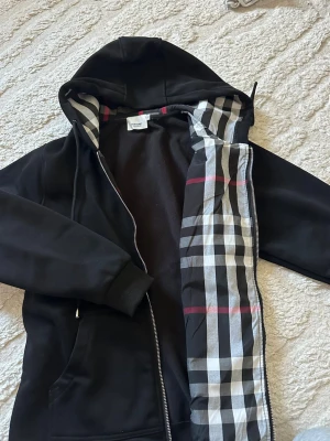 Svart hoodie med klassiskt rutmönster  - Säljer en svart hoodie från Burberry med dragkedja och huva. Insidan har det ikoniska rutmönstret i grått, vitt och rött. Hoodien har ribbade muddar och fickor framtill. Perfekt för dig som vill ha en stilren och trendig look.