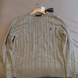 Grå kabelstickad tröja Polo Ralph Lauren - Kabelstickad grå tröja från Polo Ralph Lauren med klassisk rund hals och broderad logga på bröstet. Tröjan är i mjuk ullblandning och har ribbade muddar vid ärmslut och nederkant. Perfekt för lager-på-lager under kyliga dagar.