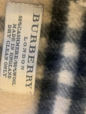 Beige rutig halsduk från Burberry - Klassisk beige halsduk från Burberry med det ikoniska rutmönstret i svart, vitt och rött. Halsduken är tillverkad i en lyxig mix av 50% kashmir och 50% ull och har fransar i ändarna. Perfekt accessoar för att lyfta din outfit.