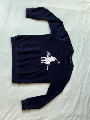 Mörkblå sweatshirt från Polo Ralph Lauren - Mörkblå sweatshirt från Polo Ralph Lauren med stor vit polospelare tryckt på bröstet. Tröjan har rund halsringning, ribbade muddar vid ärmslut och nederkant samt en avslappnad passform. Perfekt för en casual och sportig stil.