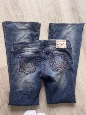 True Religion bootcut jeans blå - Snygga blå bootcut jeans från True Religion med röda kontrastsömmar och klassiska broderade bakfickor. Jeansen har en tvättad look och femficksmodell. Perfekta för dig som gillar streetstyle och vill sticka ut med detaljer.