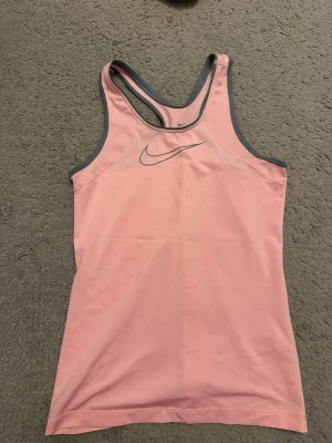 Rosa Dri-FIT linne från Nike - Sportigt rosa linne från Nike med grå kantband och Dri-FIT-material som andas. Perfekt för träning eller gympass. Snygg racerback-design och diskret Nike-logga på bröstet. Storlek 146-156. 