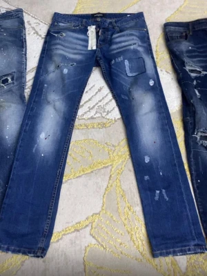  DSQUARDS Blå slitna jeans med patchdetaljer - Säljer ett par blå jeans med slitningar, patchdetaljer och färgstänk. Jeansen har en klassisk femficksmodell och raka ben. Perfekta för dig som gillar en avslappnad och trendig stil.