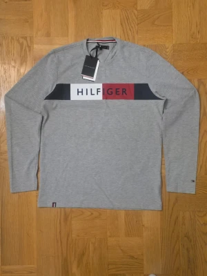 Grå långärmad tröja Tommy Hilfiger - Snygg grå långärmad tröja från Tommy Hilfiger med stort logotryck i vitt, marinblått och vinrött över bröstet. Tröjan har rund halsringning och diskret logga på ärmen. Perfekt till jeans eller chinos för en clean look.