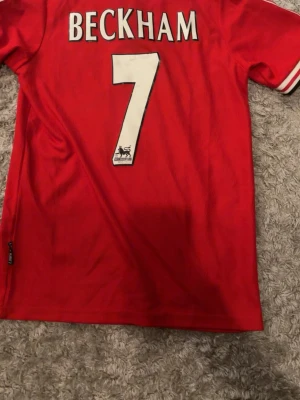 Manchester United Beckham tröja - Klassisk röd Manchester United matchtröja från Umbro med 'Beckham' och nummer 7 på ryggen. Tröjan har klubbmärke, sponsorlogga och svartvit krage med dragkedja. Perfekt för dig som älskar fotboll och retrostil.