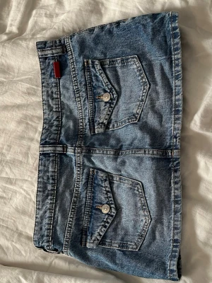 Blå jeanskjol från Divided  - Säljer en blå jeanskjol från Divided med två stora fickor framtill och knappdetaljer. Kjolen är kort och har en klassisk denimlook med ljus tvätt och synliga sömmar. Perfekt till sneakers eller boots för en cool vardagsstil.