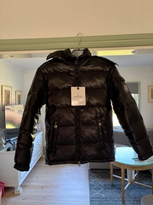 Svart dunjacka från Moncler - Snygg svart dunjacka från Moncler med glansig finish, hög krage och huva. Jackan har två dragkedjeförsedda fickor fram och Moncler-logga på ärmen. Perfekt för kalla vinterdagar och riktigt trendig stil. 