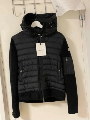 Svart pufferjacka från Moncler - Snygg svart pufferjacka från Moncler med quiltad front och huva. Jackan har ribbstickade ärmar och muddar, dragkedja framtill och fickor med tryckknapp. På vänster ärm finns en ficka med dragkedja och Moncler-logga. Perfekt för kalla vinterdagar.