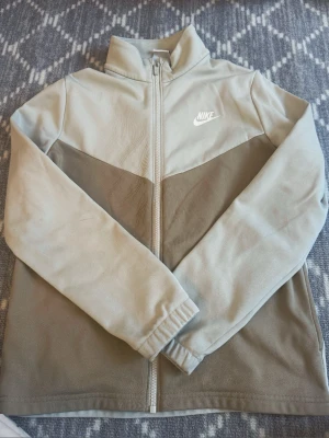 Beige vindjacka från Nike - Stilren beige vindjacka från Nike med vit logga på bröstet och ryggen. Jackan har hel dragkedja framtill, hög krage och lång ärm. Tillverkad i mjukt syntetmaterial och passar perfekt till vardagslooken.