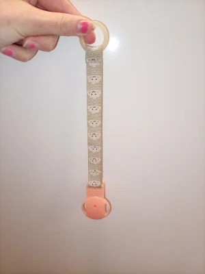 Napphållare med kattmönster - Söt napphållare med beige band dekorerat med små kattansikten och en ljusrosa plastklämma. Ringen upptill är i silikon och passar de flesta nappar. Perfekt för att hålla napp nära till hands och undvika att den tappas bort.