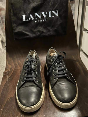 Svarta Lanvin sneakers  - Stilrena svarta sneakers från Lanvin i mjukt läder med vita sulor och svarta skosnören. Klassisk låg modell med rund tå och diskreta sömmar. Perfekta för dig som gillar minimalistisk och lyxig design.