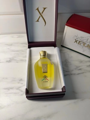 Xerjoff Naxos Eau de Parfum - Lyxig parfym från Xerjoff, Naxos, i en elegant genomskinlig glasflaska med gulddetaljer och guldfärgat lock. Flaskan har en unik form och dekoreras med en färgglad bi-symbol. Kommer i exklusiv presentförpackning med gulddetaljer.