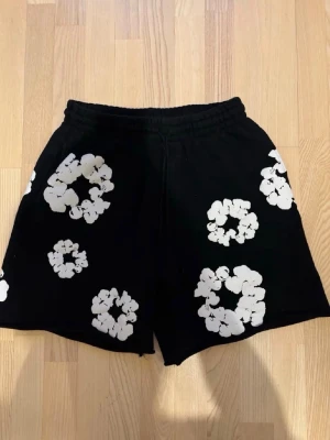 Svarta shorts med vita blommor - Snygga svarta shorts med vita blommönster i olika storlekar. Shortsen har elastisk midja för skön passform och är perfekta för varma dagar. Materialet känns mjukt och bekvämt, och designen sticker ut med sina stora, grafiska blommor.