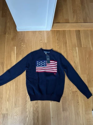 Mörkblå stickad tröja Polo Ralph Lauren - Mörkblå stickad tröja från Polo Ralph Lauren med amerikansk flagga broderad på bröstet och RL-detalj i rött. Tröjan har rund halsringning och ribbstickade muddar vid ärmslut och nederkant. Perfekt för dig som gillar klassisk och stilren design.