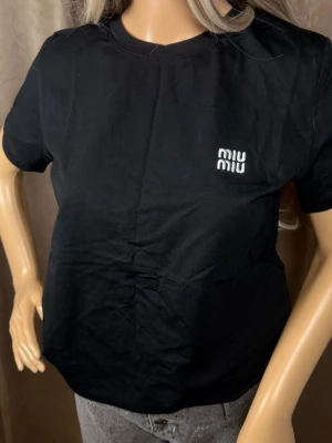 Svart Miu Miu t-shirt i bomull - Snygg svart t-shirt från Miu Miu med vit logga tryckt på bröstet. Klassisk rund halsringning och korta ärmar. Tillverkad i mjuk bomull för en bekväm känsla och enkel stil.