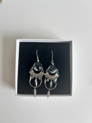 Silvriga örhängen med pärldetaljer - Unika silvriga örhängen med rund svart sten i mitten och dekorativa pärldetaljer. Hängande design med små ringar och spetsiga avslut. Snyggt boho-inspirerat mönster och fina detaljer som ger ett elegant intryck.