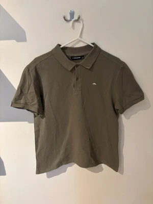 Olivgrön pikétröja från J.Lindeberg - Snygg olivgrön pikétröja från J.Lindeberg med klassisk krage och knappar framtill. Tröjan har korta ärmar och en liten vit logga på bröstet. Tillverkad i mjuk bomull som känns skön mot huden.