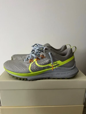 Nike React Trail grå/gul sneakers - Nike React Trail sneakers i grå mesh med neongula detaljer och reflexband. Skorna har ljusblå snören och orange öglor för extra stil. Mellansulan är dämpad och yttersulan har grovt mönster för bra grepp. Perfekta för dig som gillar streetwear och outdoor-vibe.