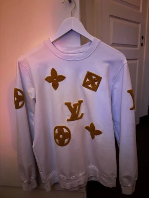 Vit Louis Vuitton sweatshirt - Vit sweatshirt från Louis Vuitton med guldiga broderade LV-loggor och monogramdetaljer på bröstet och ärmarna. Tröjan har rund halsringning, långa ärmar och ribbade muddar. Perfekt för dig som vill sticka ut med exklusiv streetwear. Sitter lite pösigt som storlek L