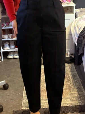 Svarta kostymbyxor med hög midja - Snygga svarta kostymbyxor med hög midja och pressveck. Byxorna har raka ben och hällor för skärp. Perfekta för en clean och stilren look. Materialet ser ut att vara en klassisk kostymblandning som ger en dressad känsla.