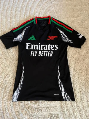 Arsenal svart matchtröja Adidas - Snygg svart Arsenal matchtröja från Adidas med gröna och röda detaljer på axlarna, vita mönster längs sidorna och tryckta loggor. Framsidan har Emirates Fly Better-tryck och Arsenal-märke, baksidan har Saliba och nummer 2. Tillverkad i lätt funktionsmaterial.