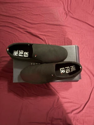 Svarta slip-on sneakers Tommy Hilfiger - Stilrena svarta slip-on sneakers från Tommy Hilfiger med diskret logga på sidan och insidan. Skorna har en låg profil, canvasmaterial och svart sula. Perfekta för en clean och avslappnad look.