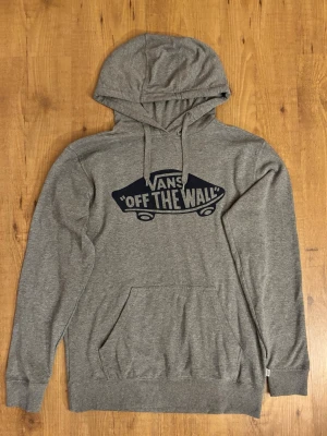Grå hoodie från Vans med tryck - Säljer en grå hoodie från Vans med klassiskt 'Off The Wall'-tryck i mörkblått på bröstet. Tröjan har huva med dragsko och en stor magficka. Materialet är mjukt och bekvämt, perfekt för chill dagar. Märke: Vans, storlek Small.