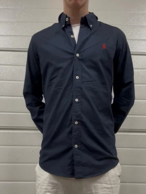 Mörkblå skjorta från Polo Ralph Lauren - Klassisk mörkblå skjorta från Polo Ralph Lauren i slim fit med vit knappstängning och röd broderad logga på bröstet. Skjortan har lång ärm och är tillverkad i mjuk bomull. Perfekt för en clean och stilren look.