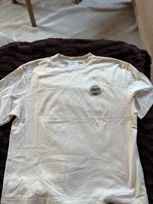 Beige kortärmad t-shirt från Arigato - Säljer en beige kortärmad t-shirt från Axel Arigato med rund hals och en liten logga på bröstet. T-shirten är i mjuk bomull och har en clean, enkel design som passar till det mesta. Perfekt basplagg för dig som gillar stilrena detaljer. Äkta såklart!