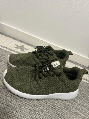 Olivgröna sneakers med vit sula från Lager 157 - Snygga olivgröna sneakers med stickad mesh-ovandel och vita, tjocka sulor. Skorna har svarta skosnören och en liten vit etikett på plösen. Perfekta för en avslappnad och trendig stil.💚