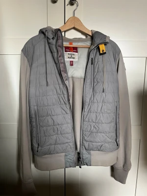 Grå pufferjacka från Parajumpers - Snygg grå pufferjacka från Parajumpers med huva och dragkedja framtill. Jackan har quiltat mönster på framsidan och axlarna, samt mjuka ärmar och rygg i sweatshirtmaterial. Två bröstfickor med dragkedja och ribbade muddar. Nypris 4000.