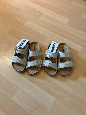 Vita sandaler från RIKKII med spännen - Snygga vita sandaler från RIKKII med två breda remmar och stora silvriga spännen. Ovandelen är i slätt material och innersulan är mjuk i mockaimitation för extra komfort. Perfekta för en avslappnad och trendig look. Köp båda för 250kr eller ett par för 150kr