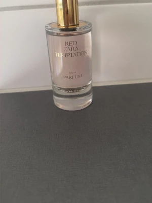 Zara Red Temptation Eau de Parfum - Zara Red Temptation Eau de Parfum med en stilren, rund glasflaska och guldig kork. Parfymen har en ljusrosa ton och elegant design med guldtext på framsidan. Perfekt för dig som gillar trendiga och lyxiga dofter.
