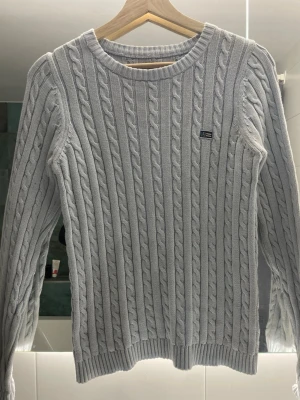 Ljusgrå kabelstickad tröja Gant - Snygg ljusgrå kabelstickad tröja från Gant med rund halsringning och diskret logga på bröstet. Tröjan har långa ärmar och ribbade muddar vid ärmslut och nederkant. Perfekt för lager på lager och klassisk stil.