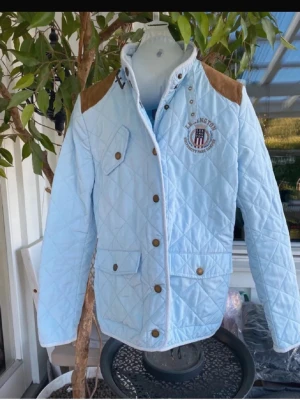 Ljusblå quiltad vindjacka Lexington - Snygg ljusblå quiltad vindjacka från Lexington med bruna mockadetaljer på axlarna och broderad logga på bröstet. Jackan har tryckknappar, hög krage och flera fickor framtill. Perfekt för dig som gillar preppy stil och vill ha något som sticker ut. Storlek M men passar bäst på S/M