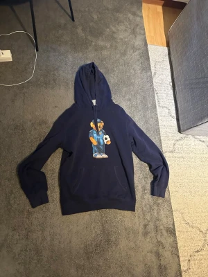 Mörkblå hoodie med nalletryck - Snygg mörkblå hoodie med huva och ett stort tryck av en nallebjörn i streetwear-stil på bröstet. Hoodien har långa ärmar, ribbade muddar och känguruficka framtill. Perfekt för dig som gillar coola och lekfulla detaljer i din outfit.