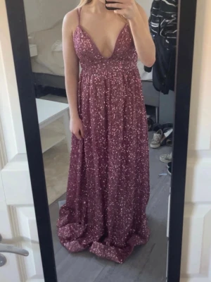 Glittrig vinröd långklänning - Säljer en vinröd långklänning med glittriga detaljer och djup v-ringning. Klänningen har smala axelband och ett flytande fall som ger en elegant siluett. Perfekt för bal eller festliga tillfällen. 
