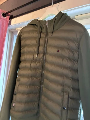 Olivgrön pufferjacka Tommy Hilfiger - Snygg olivgrön pufferjacka från Tommy Hilfiger med huva och dragkedja framtill. Jackan har quiltad framsida, mjuka ärmar och ribbade muddar. Två fickor med knappstängning och diskret logga på bröstet. Perfekt för kalla dagar. Inga defekter bortsätt från färgavtryck( sista bilden) men det är inuti luvan så ingenting som syns när man använder den 